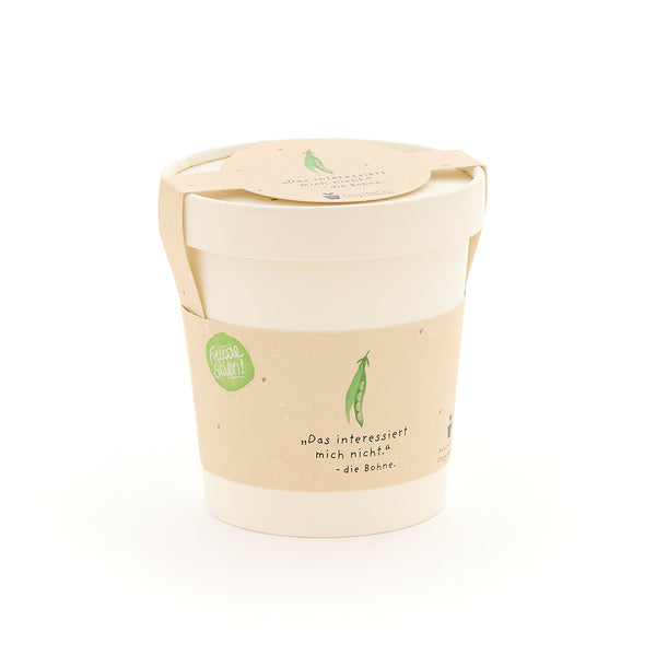 Ein weißer Becher mit beigem Etikett, auf dem eine grüne Erbse und der Spruch „Das interessiert mich nicht. – die Bohne“ abgebildet sind.