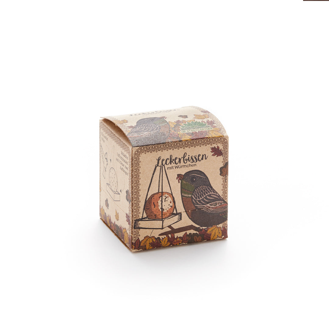 Kartonverpackung mit einer Illustration eines Vogels und eines Meisenknödels, Aufschrift "Leckerbissen mit Würmchen", herbstlich dekoriert.