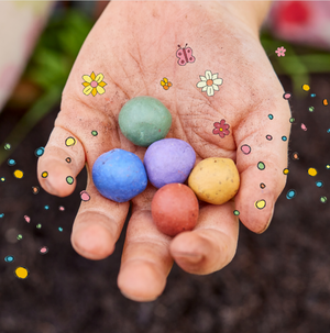 Eine Hand hält fünf bunte Samenbomben, umgeben von gezeichneten Blumen, Punkten und einem Schmetterling.