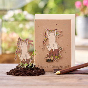 Grußkarte mit Illustration eines fröhlichen Hasen, der Wimpel hält, auf einem Baumstumpf; Text: "Hurra, der Frühling kommt!", im Vordergrund kleine Pflänzchen in Erde.