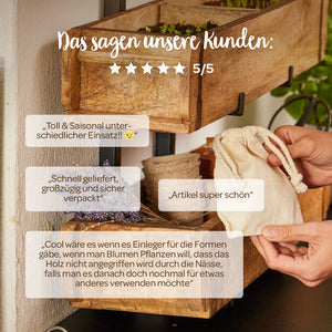 Ein Bild von einer hölzernen Pflanzenbox mit verschiedenen Kundenbewertungen auf Deutsch, eine Hand hält einen kleinen Stoffbeutel, dekorative Blumen und Pflanzen im Hintergrund.