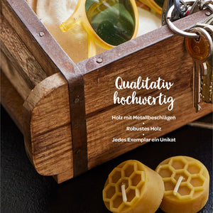 Holzkiste mit Metallbeschlägen, Sonnenbrille, Schlüsselbund und zwei Bienenwachskerzen; Text: "Qualitativ hochwertig – Holz mit Metallbeschlägen, Robustes Holz, Jedes Exemplar ein Unikat".
