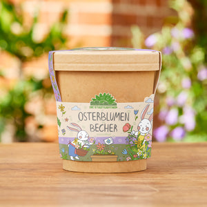 Brauner Becher mit bunter Banderole, auf der „Osterblumen im Becher“ und zwei gezeichnete Osterhasen mit Blumen abgebildet sind, im Garten auf Holzfläche stehend.