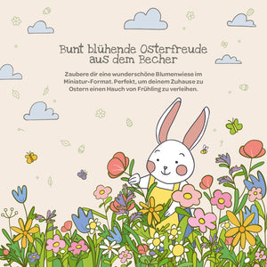 Illustration eines weißen Kaninchens in einer bunten Blumenwiese mit Frühlingsblumen, Schmetterlingen und Wolken; oben befindet sich ein deutscher Text über Osterfreude und Blumendekoration.