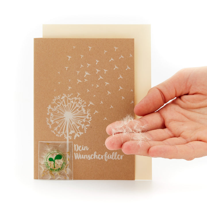 Hand hält eine Karte mit Pusteblume-Design und der Aufschrift 'Dein Wunscherfüller' sowie einer kleinen, klaren Tasche mit Pusteblumensamen"