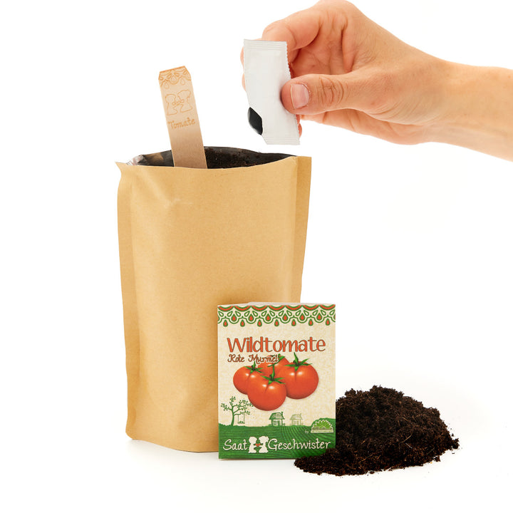 Hand fügt Samen in Anzuchterde in einer braunen Papiertüte hinzu, neben einem Samentütchen mit der Aufschrift "Wildtomate" und Erde daneben.