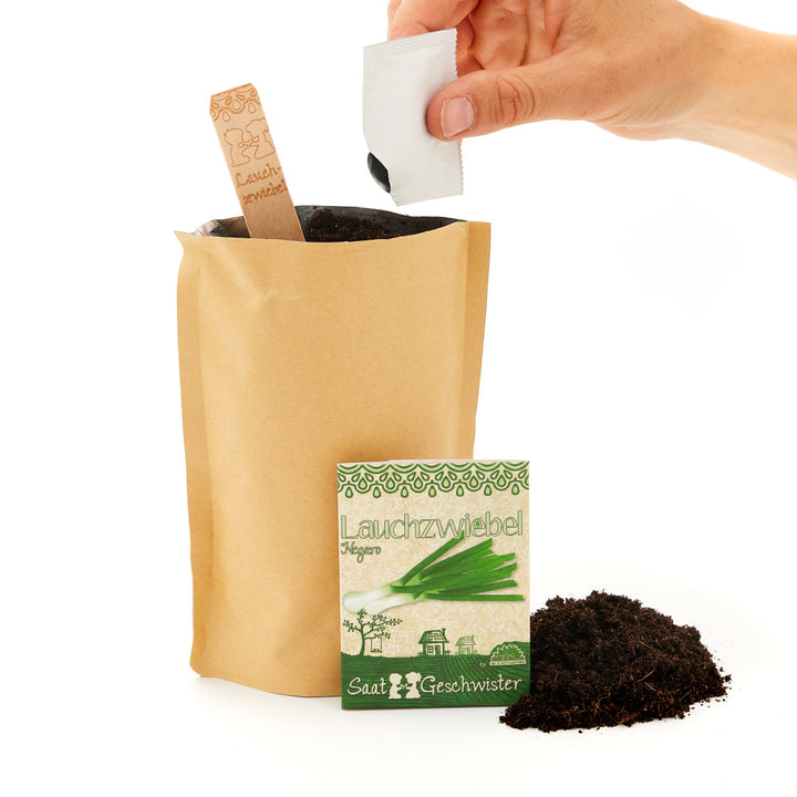 Hand hält eine Samenpackung über einen braunen Papiertüte mit Erde, neben einer weiteren Samenpackung und einem Haufen Erde.