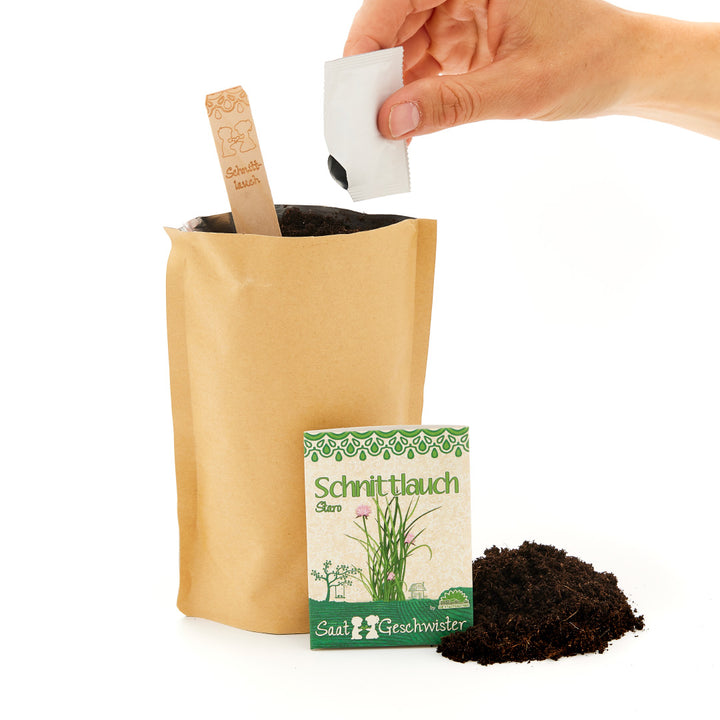 Eine Hand, die ein Samentütchen über einem Kraftpapierbeutel mit Erde hält, daneben eine Packung mit der Beschriftung "Schnittlauch" und ein Haufen Erde.