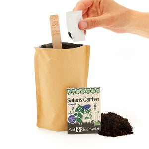 Eine Hand, die einen Samenbeutel über eine Papiertüte mit Erde hält, neben der ein Schild mit der Aufschrift "Satans Garten" und ein weiterer Samenbeutel stehen.
