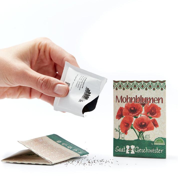 Eine Hand, die ein Samenpäckchen hält, neben einem zweiten Päckchen mit der Aufschrift "Mohnblumen" und einigen verstreuten Samen auf einer weißen Oberfläche.