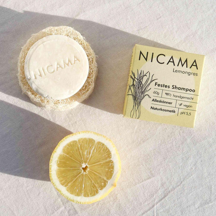 Festes Shampoo von NICAMA mit Zitronengrasduft neben einer Zitronenscheibe und der Verpackung auf einer hellen Oberfläche.