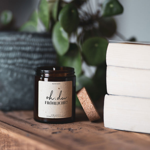 Eine Duftkerze mit der Aufschrift "oh, du Fröhliche!" neben einem Stapel Bücher auf einem Holztisch mit unscharfen Pflanzen im Hintergrund.