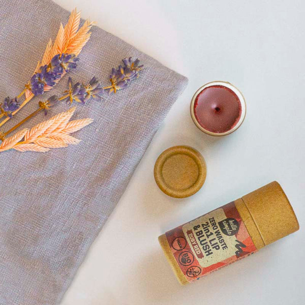 Stilleben mit getrockneten Lavendelzweigen, einer roten Kerze und einer runden Pappverpackung auf einem grauen Stoff.