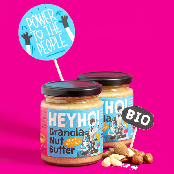 Zwei Gläser Bio-Nussbutter mit der Aufschrift "HEYHO! Granola Nut Butter", umgeben von Nüssen auf einem pinken Hintergrund, mit einem Schild "POWER TO THE PEOPLE" und einem "BIO"-Logo.