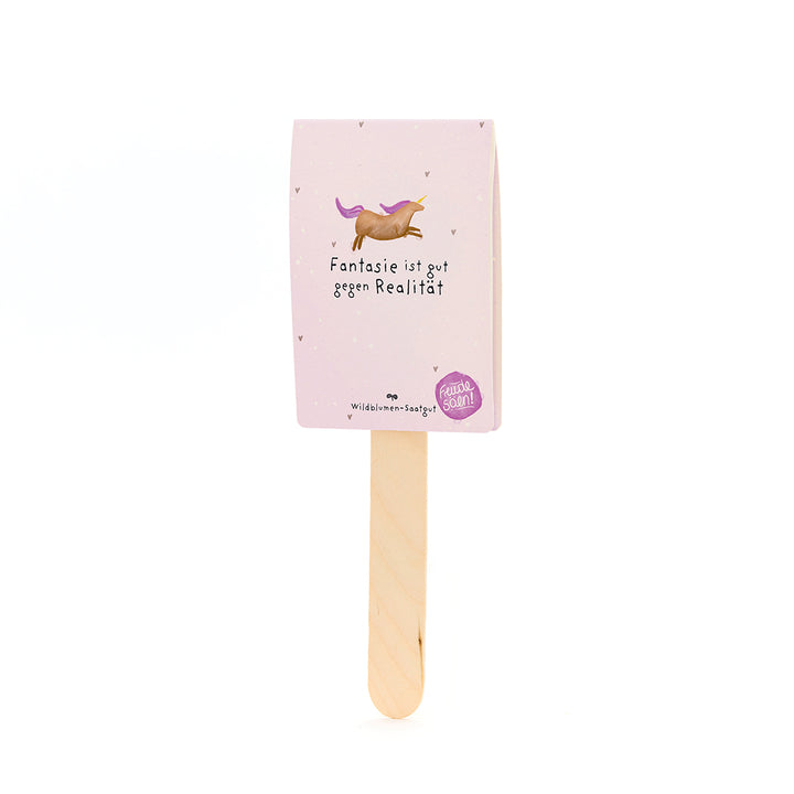 Holzstab mit rosa Karte, darauf ein gezeichnetes Einhorn und der Text "Fantasie ist gut gegen Realität", darunter steht "Wildblumen-Saatgut".