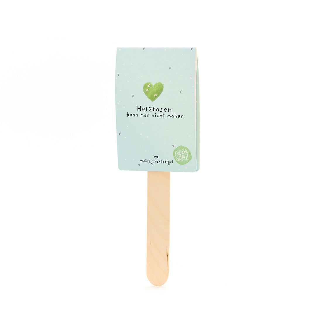 Holzstab mit hellblauem Schild, auf dem ein grünes Herz und der Text "Herzrasen kann man nicht mähen" sowie "Weidegras-Saatgut" steht.