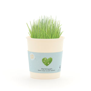 Becher mit grünem Gras und einer Banderole, auf der ein grünes Herz und der Text „Herzrasen kann man nicht mähen“ abgebildet sind.