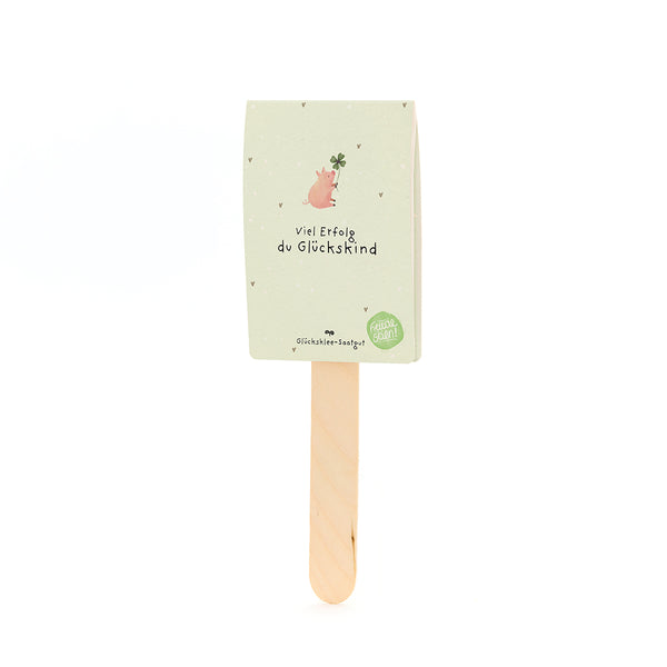 Ein Holzstab mit einer Karte, auf der ein Schwein mit Kleeblatt und der Text „Viel Erfolg du Glückskind“ steht.