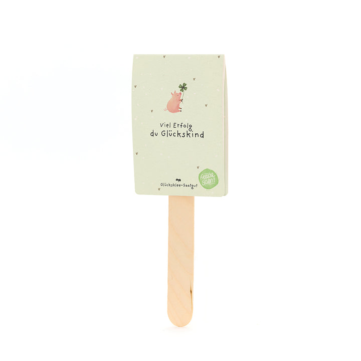 Ein Holzstab mit einer Karte, auf der ein Schwein mit Kleeblatt und der Text „Viel Erfolg du Glückskind“ steht.