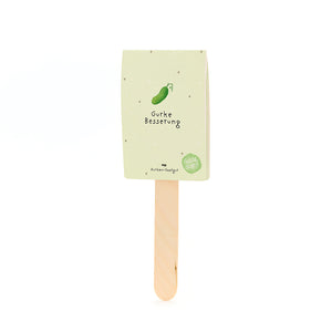 Ein Holzstab mit einem beige-grünen Papieretikett, auf dem eine gezeichnete Gurke und der Text "Gurke Besserung" zu sehen sind.