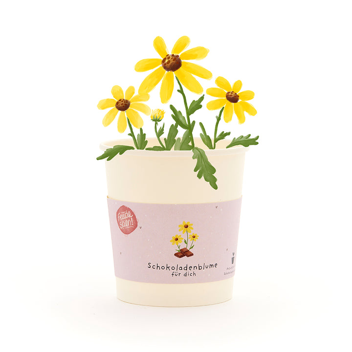 Becher mit gelben Schokoladenblumen und grünem Blattwerk, Etikett mit Aufschrift „Schokoladenblume für dich“.