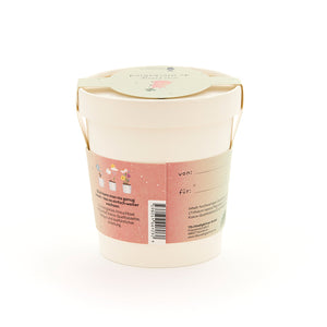 Beiger Blumentopf mit Henkel, rosa Etikett mit Pflanzillustrationen und Text auf Deutsch, Geschenkanhänger auf dem Deckel.