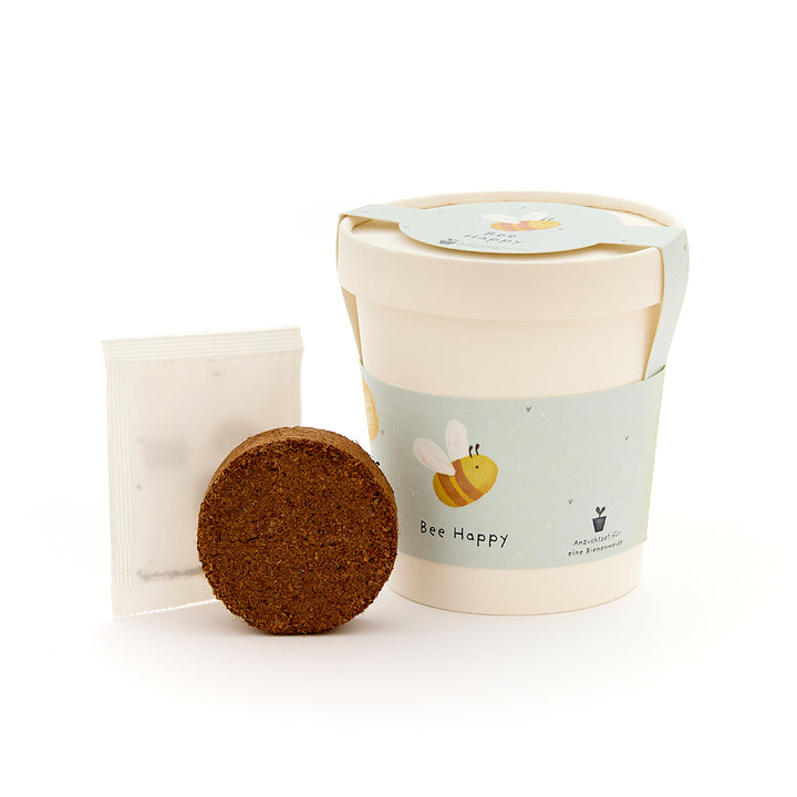 Pflanzset mit rundem Quelltablette, Samenpäckchen und einem Becher mit Bienenmotiv und der Aufschrift "Bee Happy".