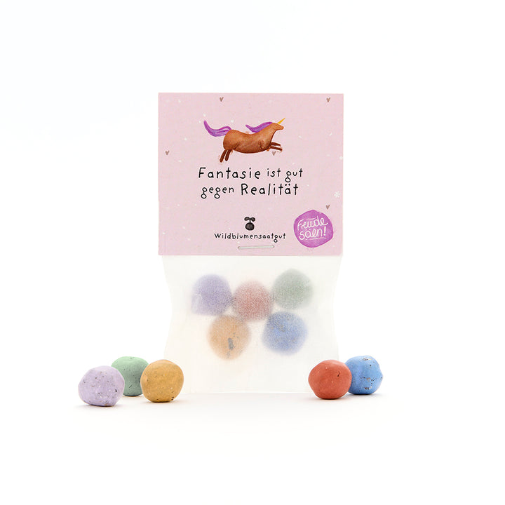 Tüte mit bunten Samenbällen und einer lilafarbenen Verpackung mit Einhorn und dem Text „Fantasie ist gut gegen Realität“.