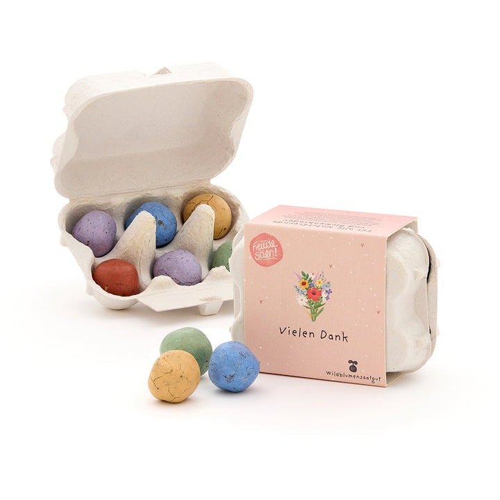 Sechs bunte Samenbomben in einer kleinen Eierkarton-Verpackung, daneben eine geschlossene Box mit der Aufschrift "Vielen Dank" und einer Blumenillustration.