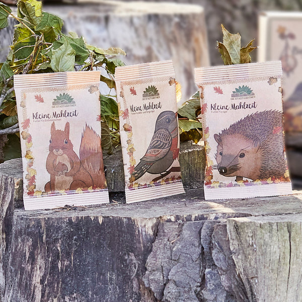 Drei Packungen für Tierfutter mit Abbildungen eines Eichhörnchens, eines Vogels und eines Igels auf Baumstämmen"