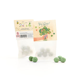 Tüte mit grünen Saatkugeln und Glücksklee-Samen, daneben mehrere lose grüne Kugeln, auf der Verpackung Glückwunschmotiv und Kleeillustration.