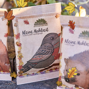 Papiertüte mit Aufdruck eines Vogels und der Aufschrift "Kleine Mahlzeit – Futter für Wild- und Singvögel" von Die Stadtgärtner, umgeben von herbstlichen Blättern.