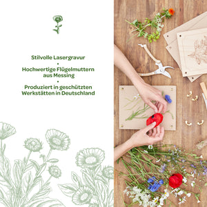 Zwei Hände arrangieren bunte Blumen auf einem Holzrahmen; daneben liegen Werkzeuge, Messing-Flügelmuttern und gravierte Holzplatten auf einem Holztisch. Links Text mit Informationen über stilvolle Lasergravur und Produktion in Deutschland.