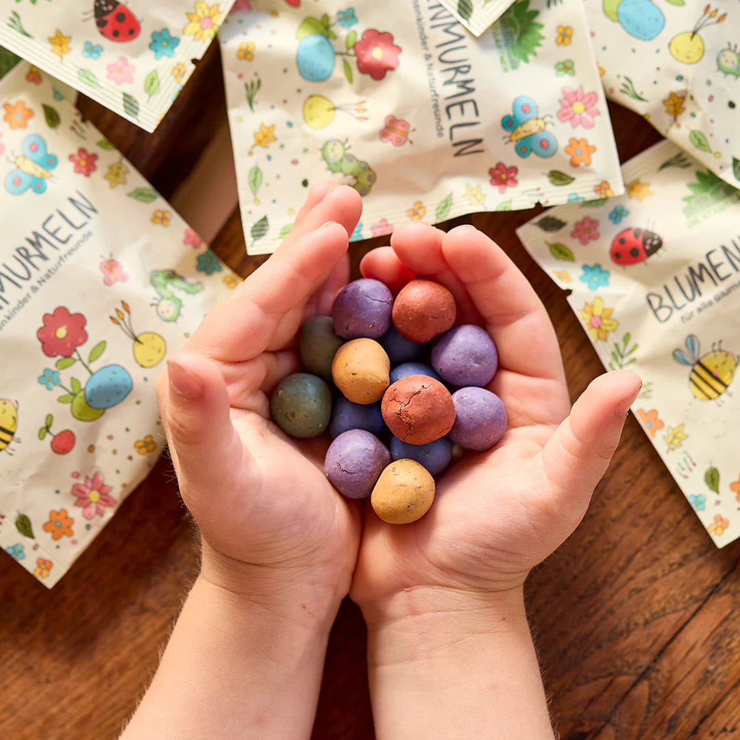 Kinderhände halten bunte Samenbomben, im Hintergrund liegen bunt bedruckte Verpackungen mit Blumen- und Insektenmotiven.