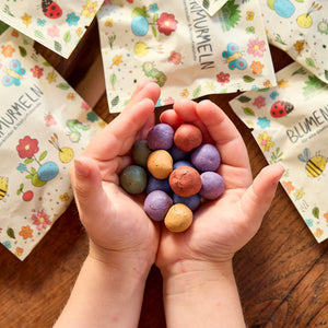 Kinderhände halten bunte Samenbomben, im Hintergrund liegen bunt bedruckte Verpackungen mit Blumen- und Insektenmotiven.
