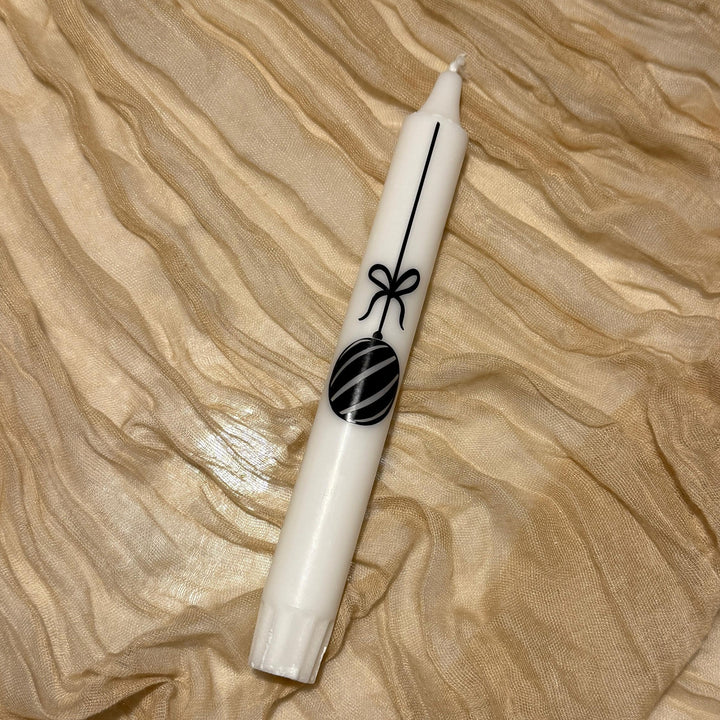 Eine weiße Stabkerze mit einer schwarzen Weihnachtskugel als Design liegt auf einer goldenen Stoffbordüre