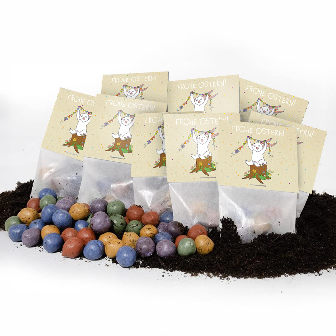 Bunte Samenbomben liegen auf Erde, dahinter mehrere Tütchen mit Aufschrift „Frohe Ostern!“ und einem Hasen-Motiv.