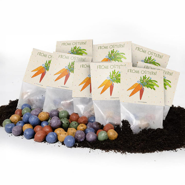 Mehrere Tütchen mit der Aufschrift „Frohe Ostern!“ und einer Karottenzeichnung, davor bunte Samenbälle und Erde.
