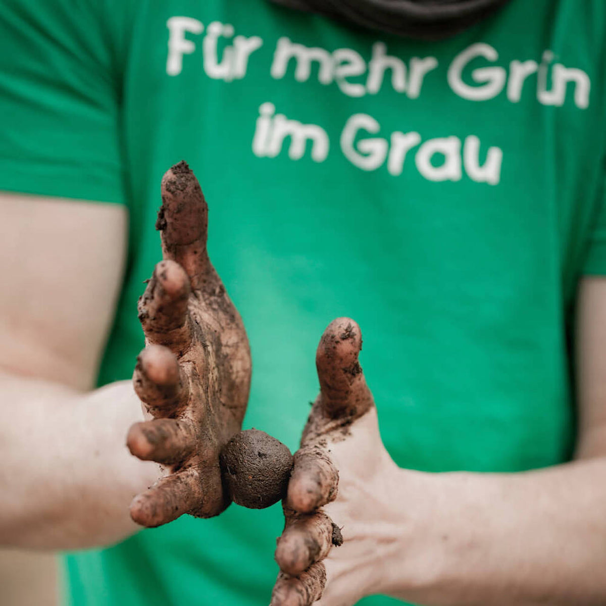 Person in grünem T-Shirt mit der Aufschrift "Für mehr Grün im Grau" hält verschmutzte Hände vor die Kamera.