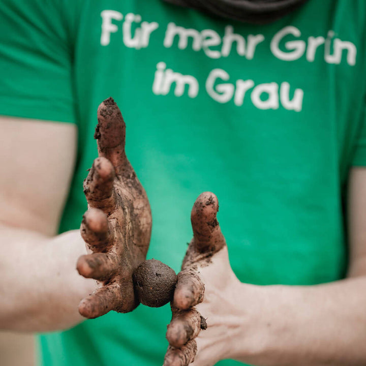 Person zeigt verschmutzte Hände vor grünem T-Shirt mit Aufschrift "Für mehr Grün im Grau"