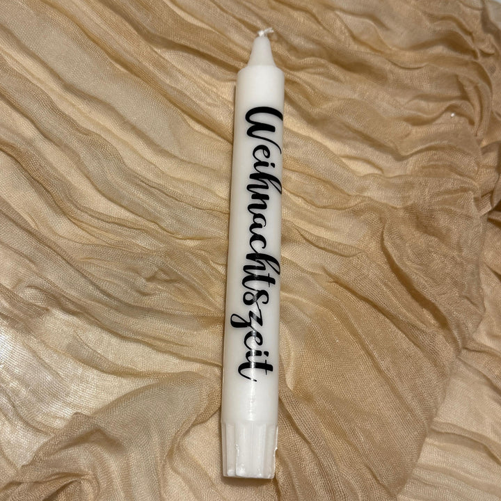 Weiße Stabkerze mit dem schwarzen Schriftzug "Weihnachtszeit" an der Seite auf einem goldenen Stoff drapiert