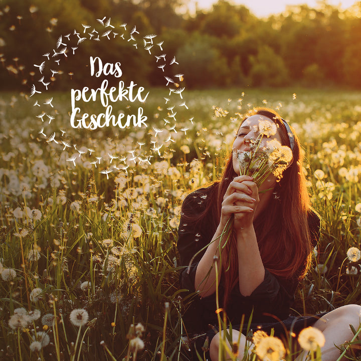 Frau sitzt in einem Feld voller Löwenzahn bei Sonnenuntergang und riecht an Blumen mit Text "Das perfekte Geschenk".