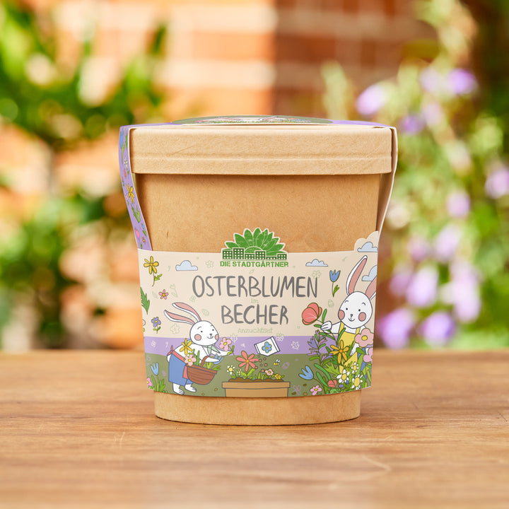 Brauner Becher mit bunter Banderole, auf der „Osterblumen im Becher“ und zwei gezeichnete Osterhasen mit Blumen abgebildet sind, im Garten auf Holzfläche stehend.