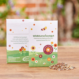 Zwei Packungen Wildblumensaatgut mit bunter Blumenillustration und ein kleiner Haufen Saatgut auf einem Holztisch im Freien.