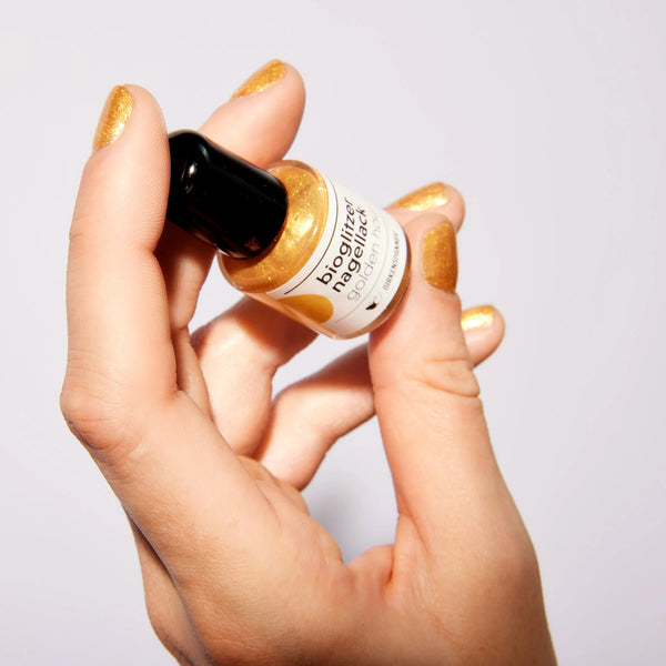 Hand hält eine Flasche Nagellack mit goldenem Glitzerfinish auf den Nägeln.