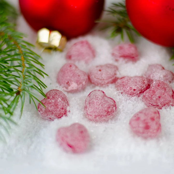 Rote herzförmige Süßigkeiten im Schnee mit Tannenzweig und Weihnachtsdekoration im Hintergrund.