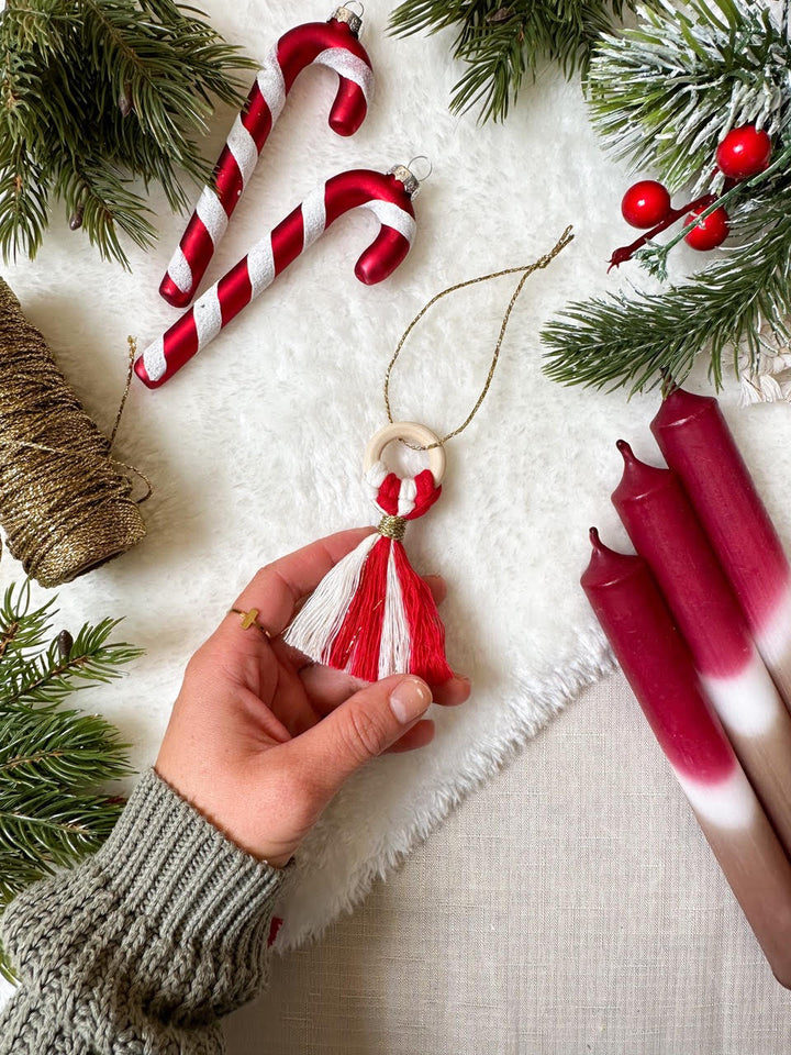 Hand hält selbstgemachten Weihnachtsschmuck neben festlicher Dekoration mit Tannenzweigen und Kerzen.