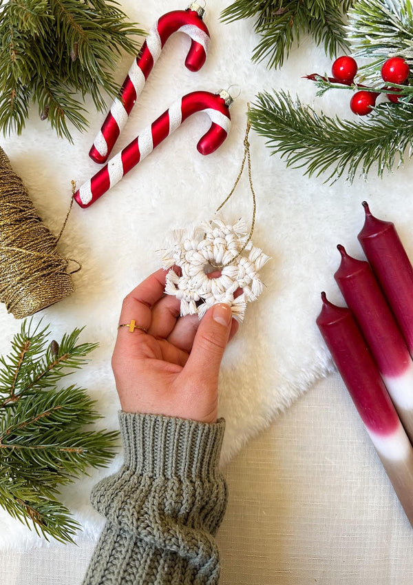 Hand hält eine weiße Weihnachtsbaumdekoration mit Tannenzweigen, Zuckerstangen, einer Garnrolle und Kerzen im Hintergrund.