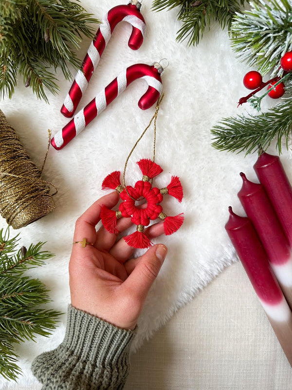 Eine Hand hält eine rote Weihnachtsdekoration, umgeben von Zuckerstangen, grünen Tannenzweigen und roten Kerzen auf einem weißen Untergrund.