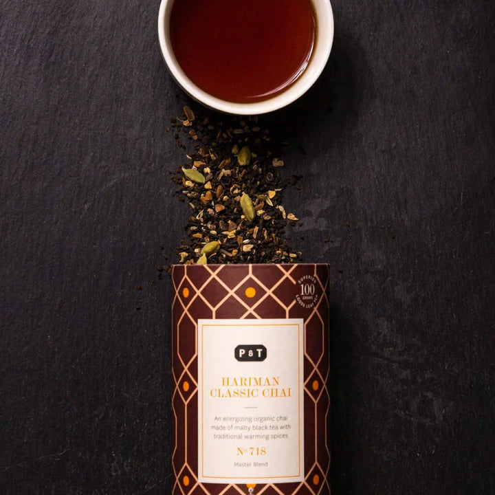 Eine Tasse Chai-Tee neben einer offenen Teepackung mit losen Teeblättern und Gewürzen auf einer dunklen Oberfläche.